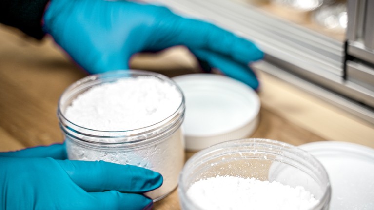Blue nitrile-gloved hands holding an open jar filled with a clumpy white powder