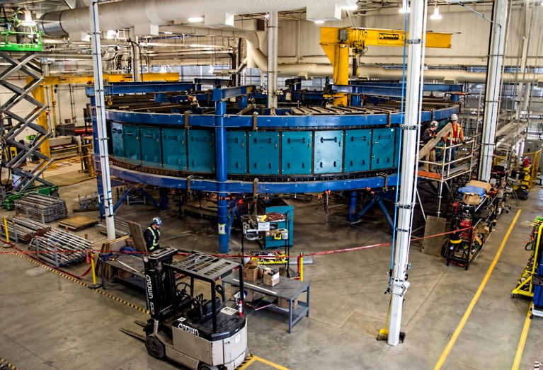 In a large open factory space, a blue, 14-metre wide wheel-like carbon capture machine stands horizontal to the ground, elevated by a blue frame